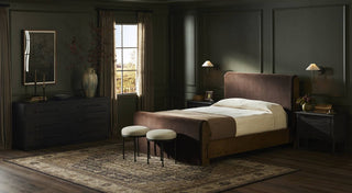 A moody modern bedroom featuring a dark green paneled wall, a plush upholstered bed with a brown velvet headboard, and two hammered iron accent stools with round upholstered seats at the foot of the bed.